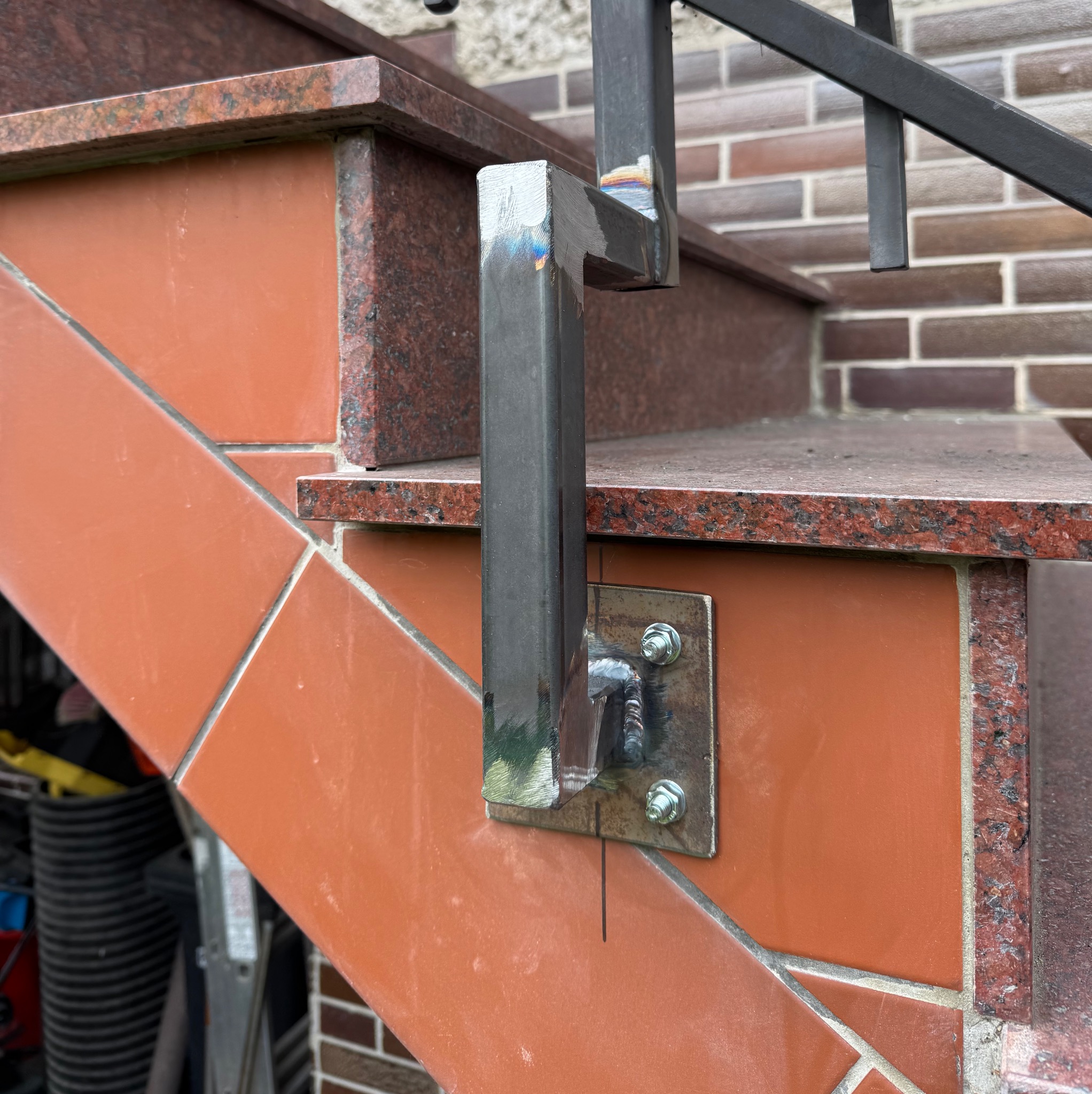 Spawana konstrukcja metalowa, mocowana do schodów z terakoty i granitu. Widoczne spawy i śruby mocujące. Element balustrady w trakcie montażu.