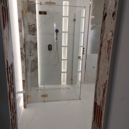 PAT House - Nowoczesna kabina prysznicowa z marmurowymi ścianami i złotymi detalami, widoczna przez niedokończone futryny drzwiowe w Rzeszowie. Biała marmurowa podłoga i oświetlenie LED podkreślają elegancję.