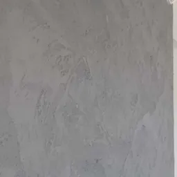 Szara ściana pokryta dekoracyjną, nierówną strukturą tynku, z widocznym fragmentem białego sufitu i kablem.