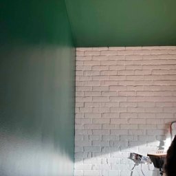PAT House - Fragment wnętrza z zielonym, świeżo pomalowanym sufitem i ścianą oraz białą ścianą z imitacji cegły; widoczne gniazdka elektryczne i lampa budowlana.
