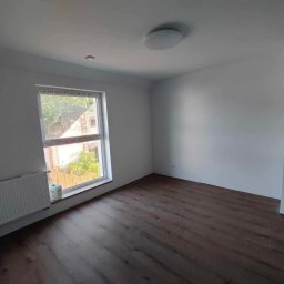 PAT House - Pusty pokój po remoncie z panelami podłogowymi w odcieniach brązu, białymi ścianami, grzejnikiem pod oknem i okrągłą lampą sufitową.