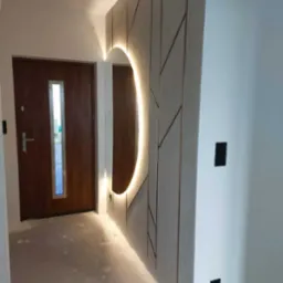 Nowoczesne wykończenie przedpokoju z podświetlanym lustrem i dekoracyjną ścianą. Brązowe drzwi wejściowe z przeszkleniem. Szary surowy beton na podłodze.