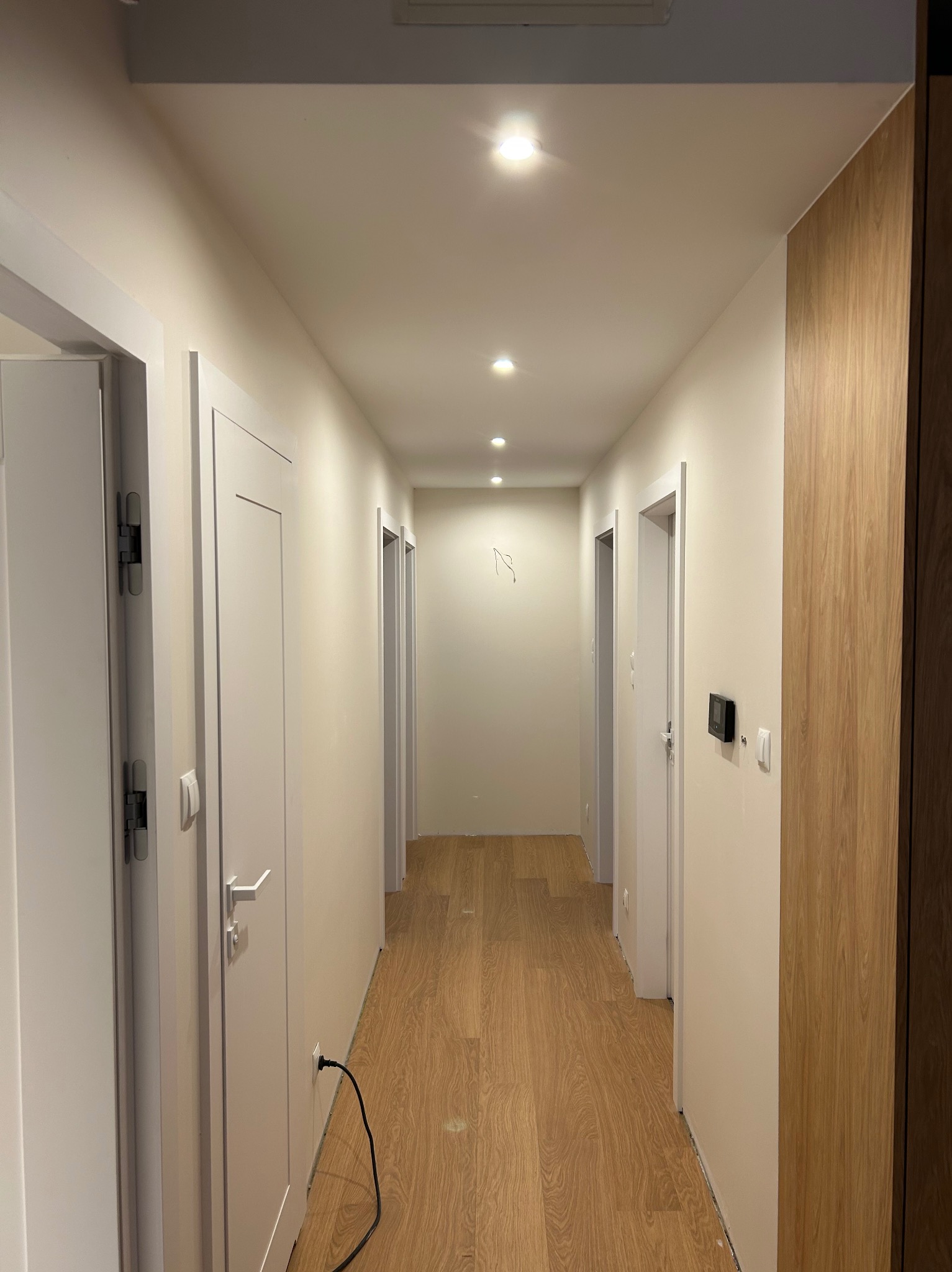 Długi korytarz po remoncie z drewnianą podłogą, białymi drzwiami i świeżo pomalowanymi ścianami w odcieniach beżu, widoczne gniazdko elektryczne i kabel przy jednej ze ścian.