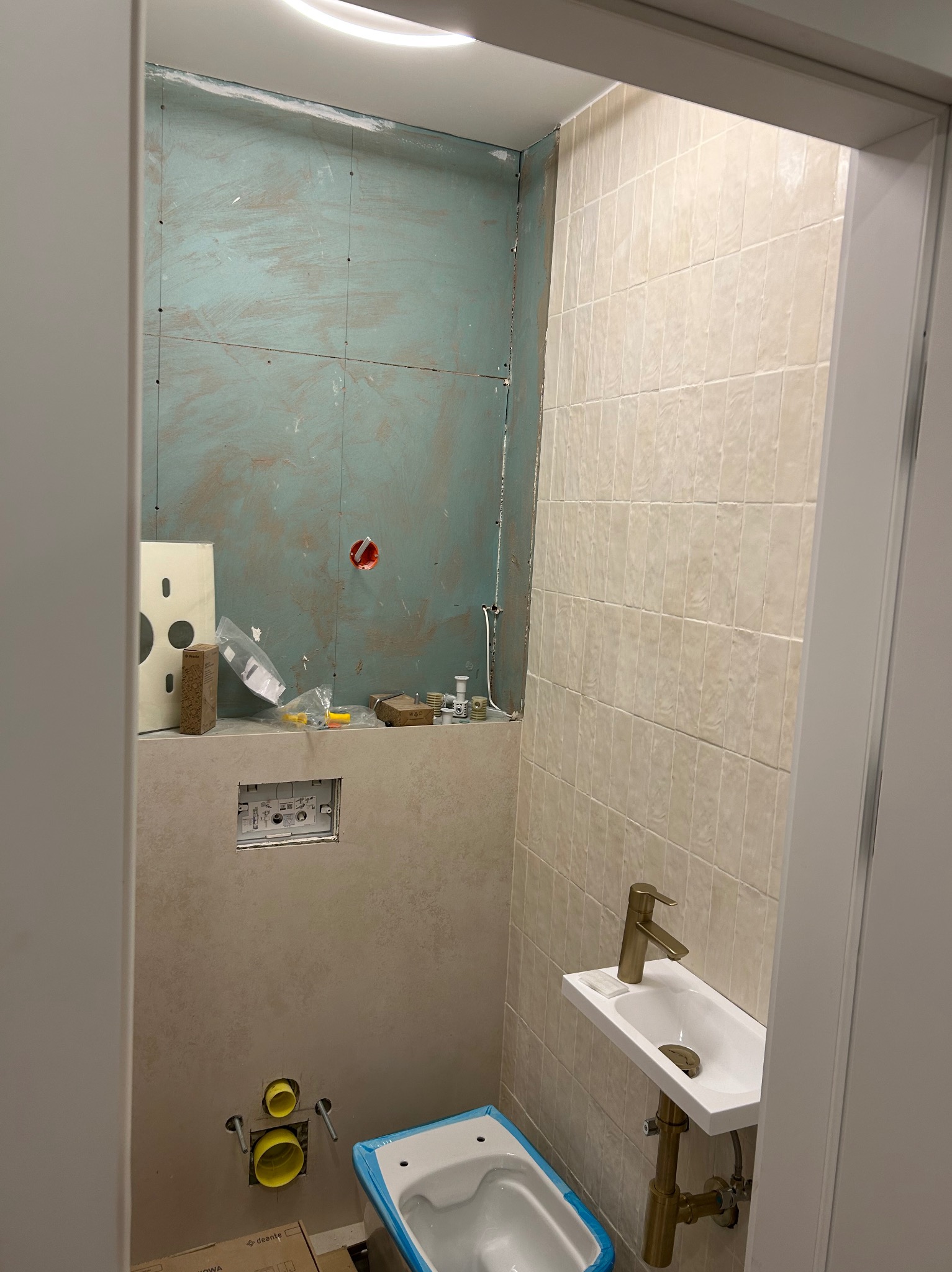 Remont łazienki w trakcie: widok na częściowo wykończoną ścianę z zielonymi płytami gipsowymi, przygotowane instalacje pod toaletę, zamontowana umywalka z baterią w kolorze szczotkowanego złota...