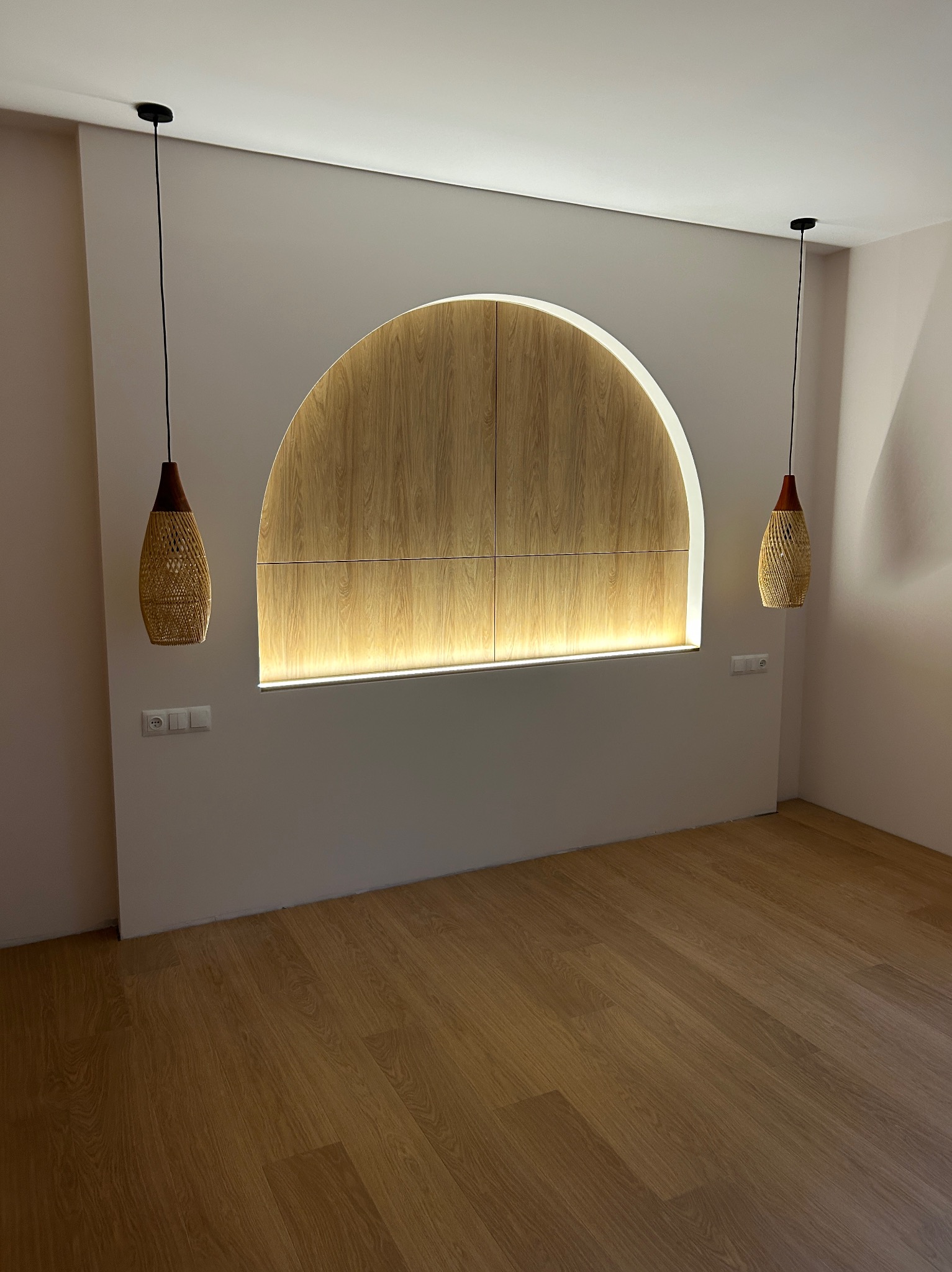 Wnęka w kształcie łuku z drewnianym wykończeniem i podświetleniem LED, wiszące lampy z abażurami z plecionej rafii, panele podłogowe o naturalnym odcieniu drewna.