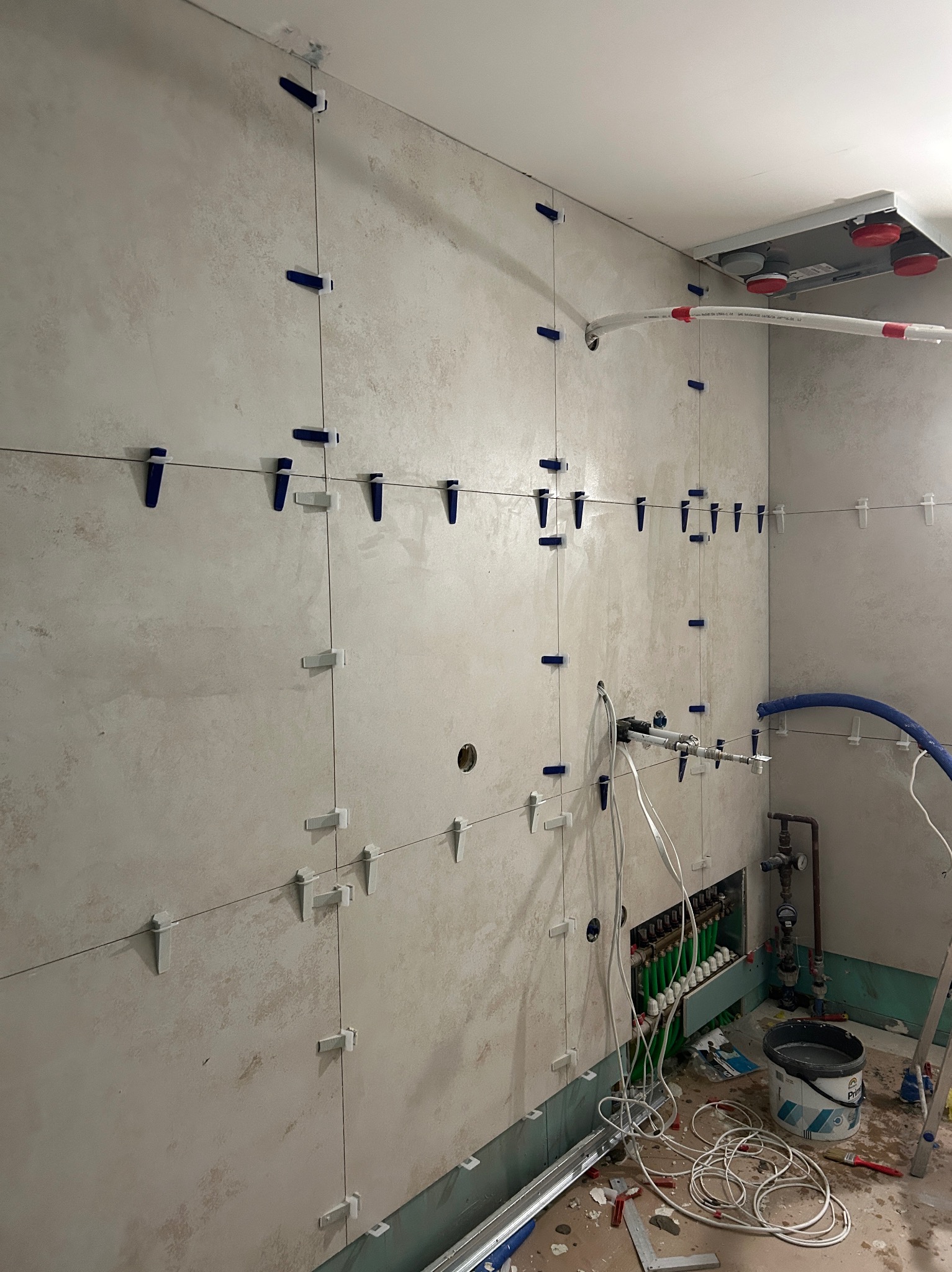Ściana w trakcie układania płytek ceramicznych, z użyciem klipsów poziomujących, widoczne instalacje wodne i grzewcze, oraz narzędzia pracy na podłodze.