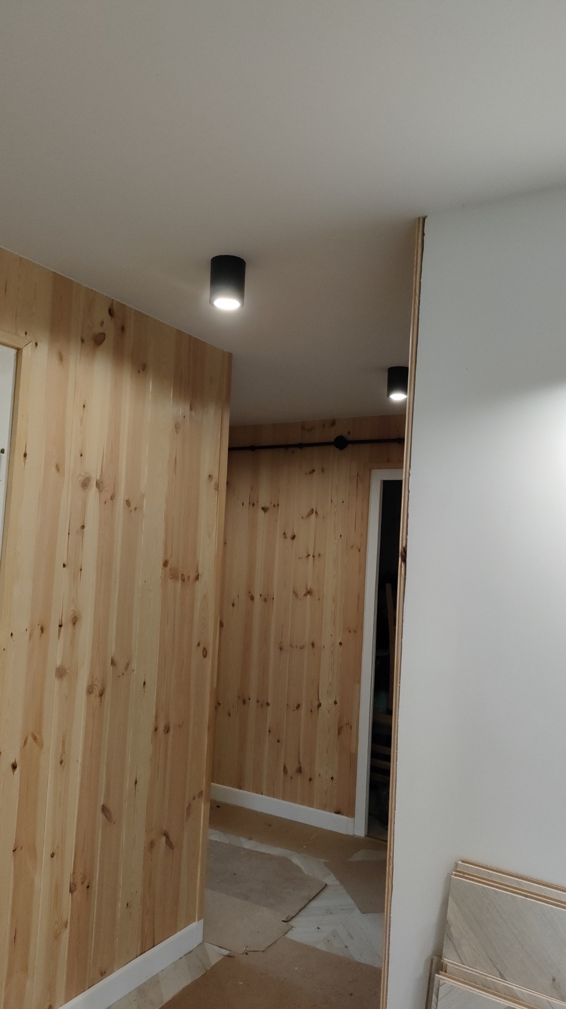 Wnętrze w trakcie prac wykończeniowych: ściany wyłożone jasnym drewnem, sufit z zamontowanymi czarnymi lampami punktowymi, widoczne materiały podłogowe przygotowane do montażu.