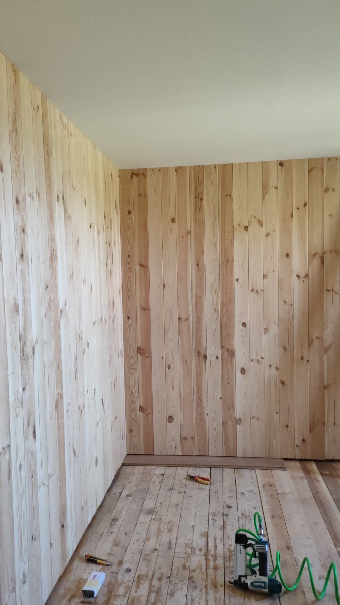 Pokój w trakcie wykańczania boazerią drewnianą, widoczne narzędzia: zszywacz pneumatyczny, obcęgi i pudełko ze zszywkami.