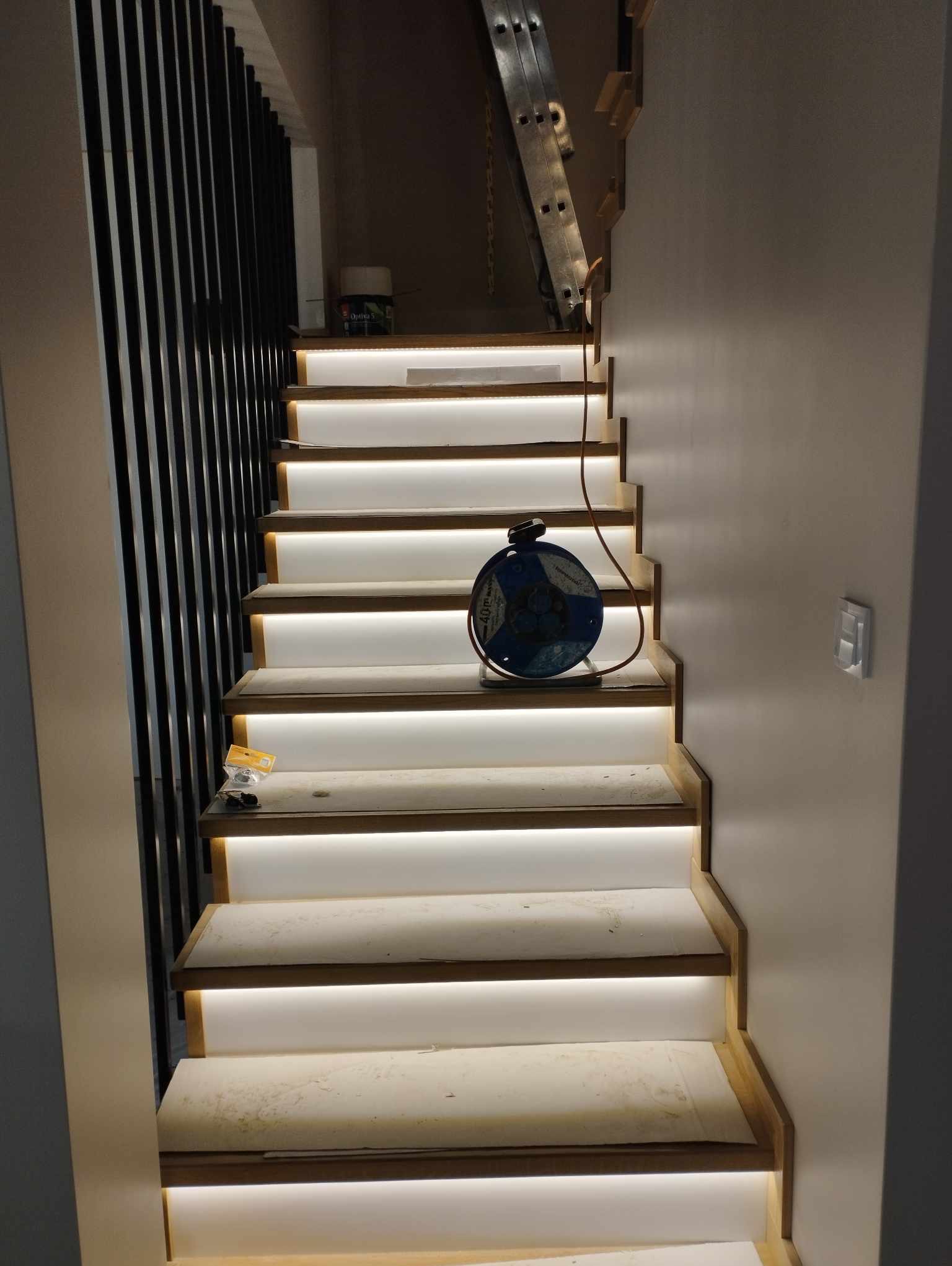 Schody z podświetleniem LED w trakcie montażu, widoczna drabina, barierka z czarnych listew, kabel na schodach i narzędzia.