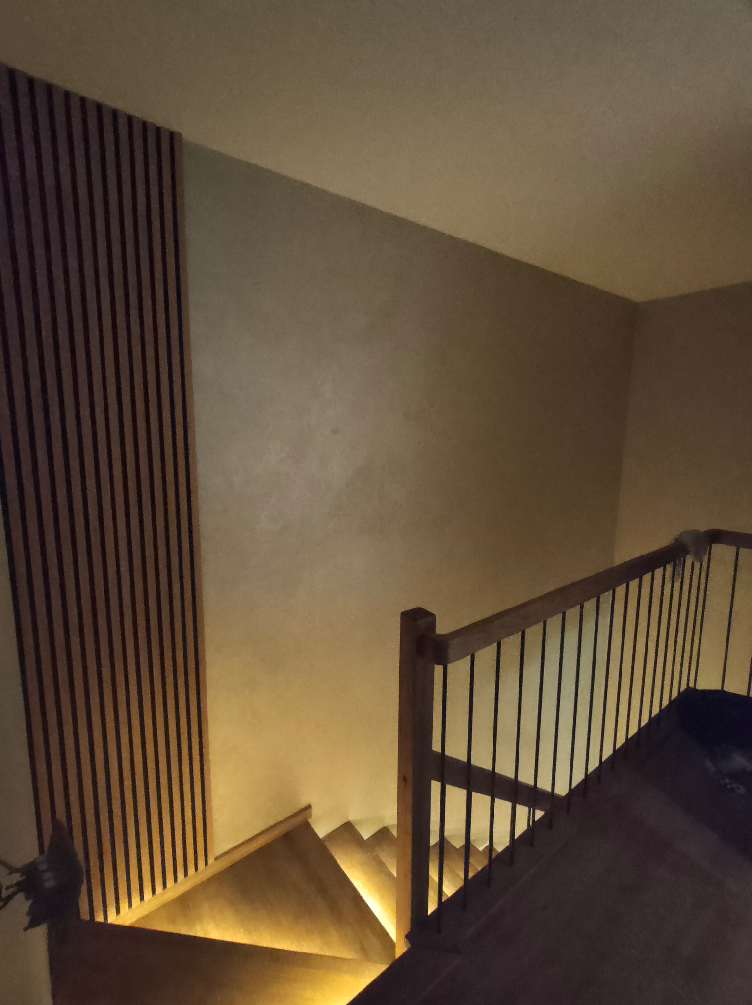 Klatka schodowa z drewnianymi schodami podświetlonymi od dołu, balustrada z drewnianymi słupkami i czarnymi, pionowymi prętami, dekoracyjna ściana z pionowych listew.