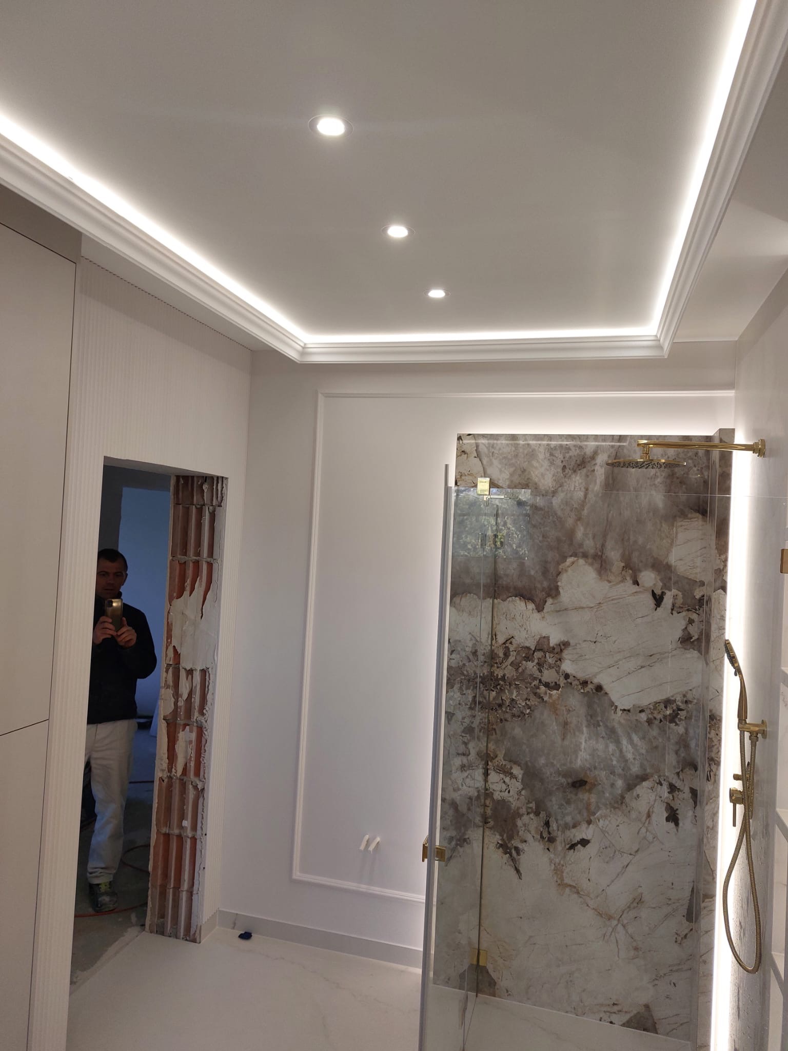 Wykończona łazienka z kabiną prysznicową z kamienną ścianą, oświetleniem LED na suficie i pracownikiem robiącym zdjęcie w sąsiednim pomieszczeniu.