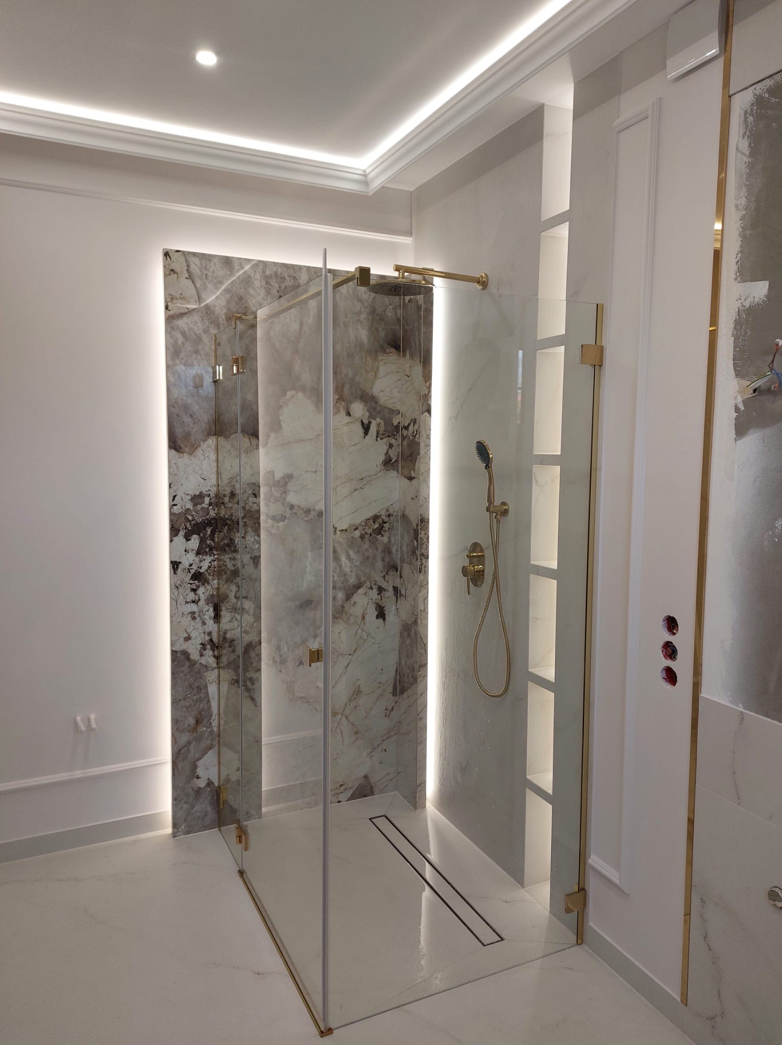 Nowoczesna kabina prysznicowa ze złotymi detalami, marmurowym wykończeniem ścian i podświetleniem LED, widoczne przygotowane otwory na gniazdka elektryczne.