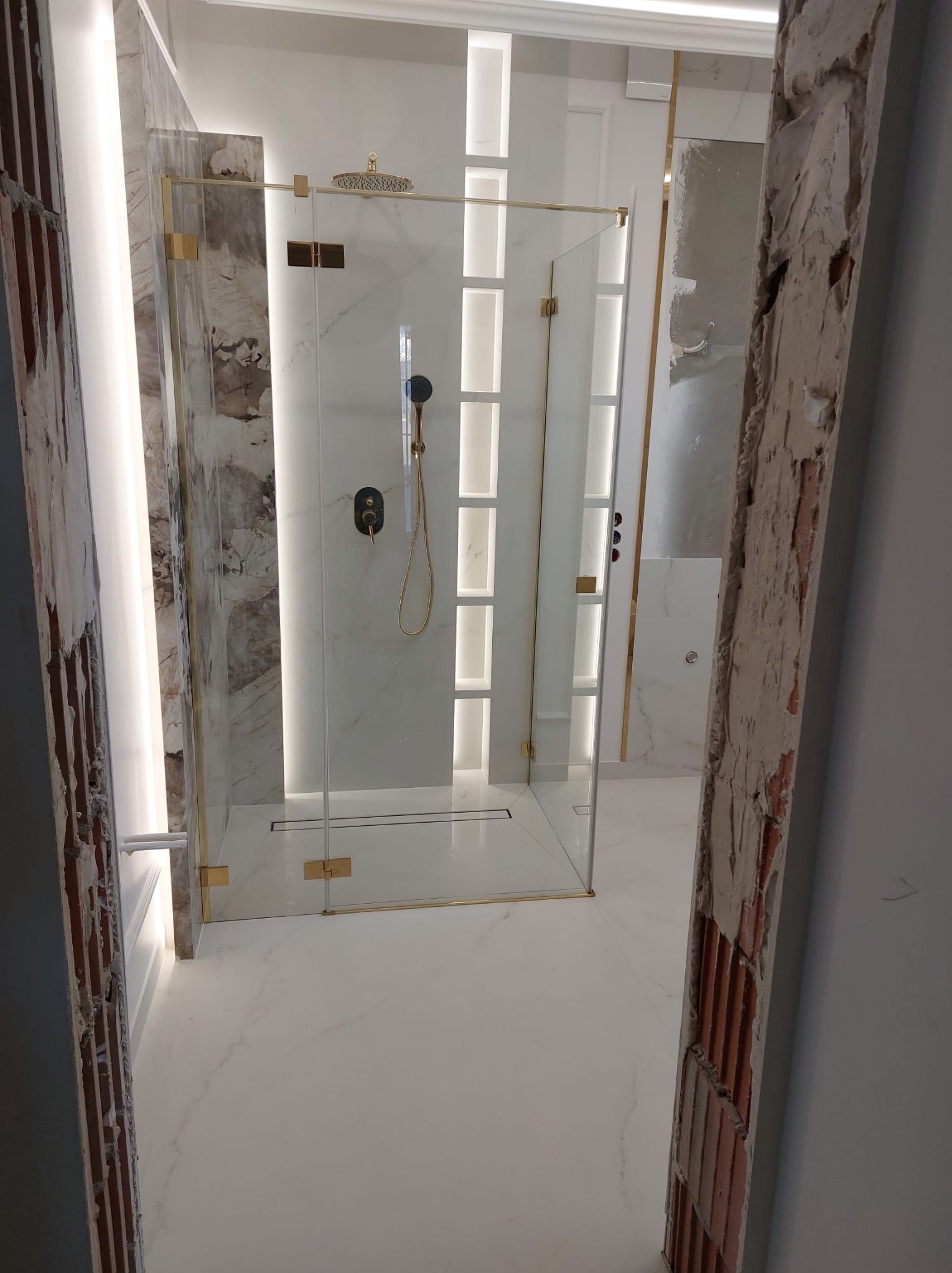 Nowoczesna kabina prysznicowa z marmurowymi ścianami i złotymi detalami, widoczna przez niedokończone futryny drzwiowe w Rzeszowie. Biała marmurowa podłoga i oświetlenie LED podkreślają elegancję.