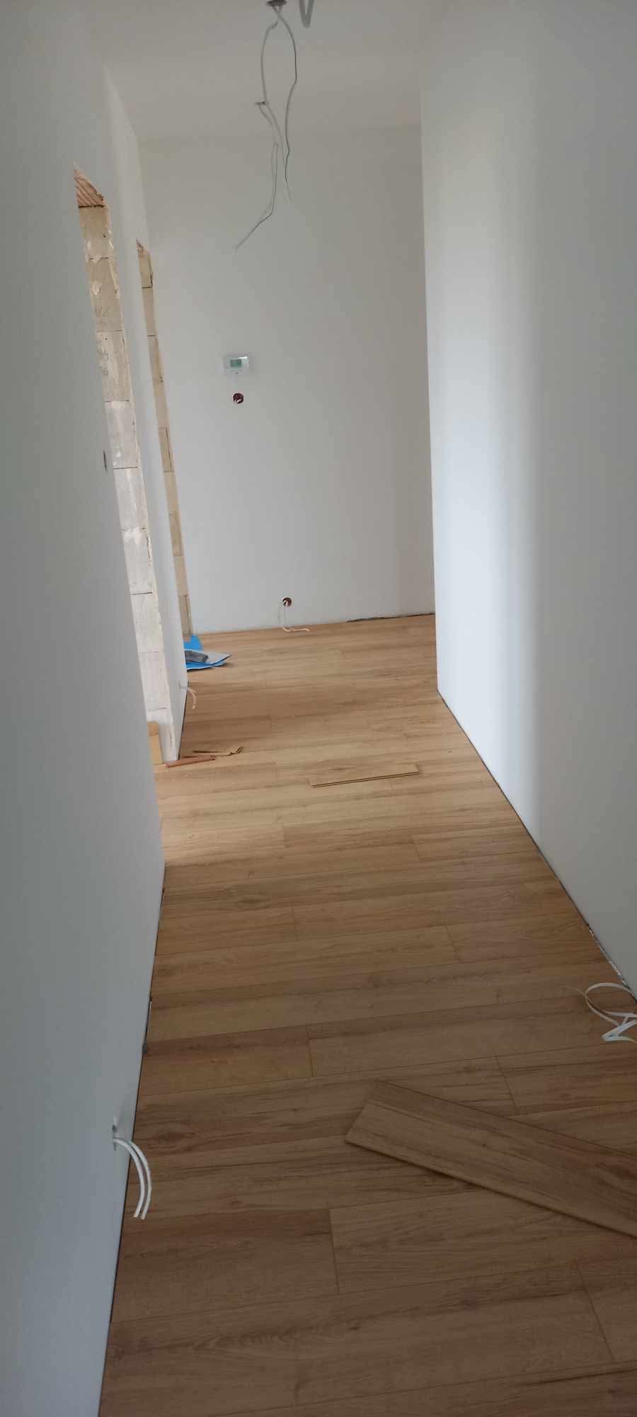 Korytarz w trakcie wykańczania z położoną drewnopodobną podłogą, widoczne wystające kable ze ściany i luźne panele podłogowe.