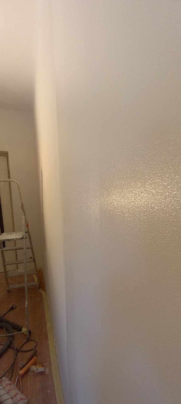 Świeżo pomalowana ściana w jasnym odcieniu z widoczną fakturą, drabina malarska oparta o ścianę, narzędzia malarskie na podłodze, widoczne ślady po taśmie malarskiej.