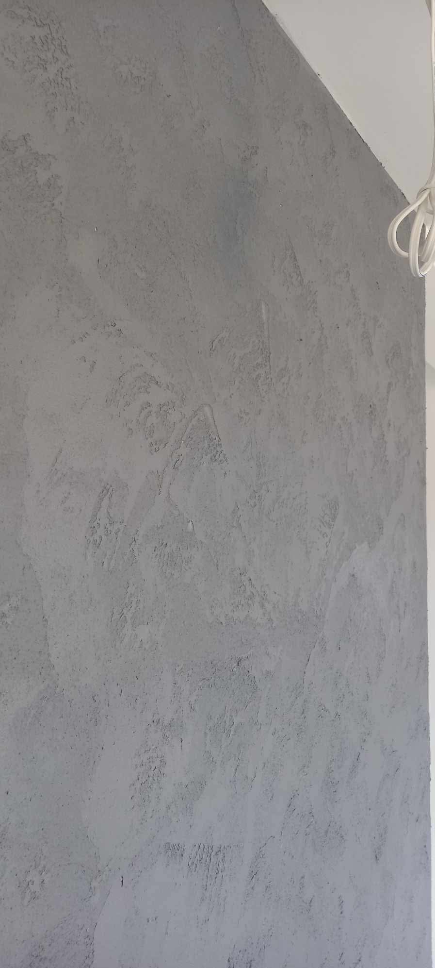 Szara ściana pokryta dekoracyjną, nierówną strukturą tynku, z widocznym fragmentem białego sufitu i kablem.
