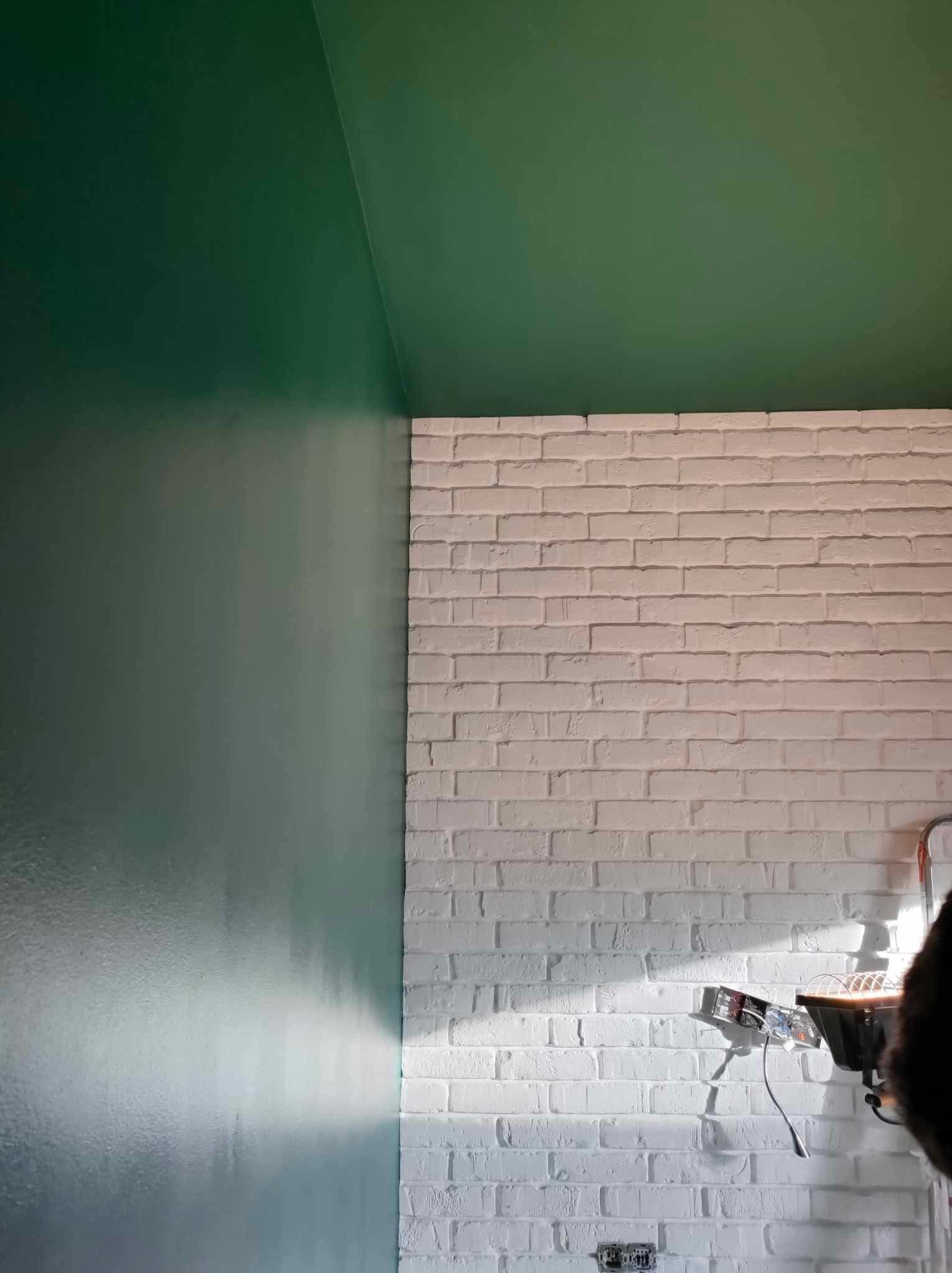 Fragment wnętrza z zielonym, świeżo pomalowanym sufitem i ścianą oraz białą ścianą z imitacji cegły; widoczne gniazdka elektryczne i lampa budowlana.