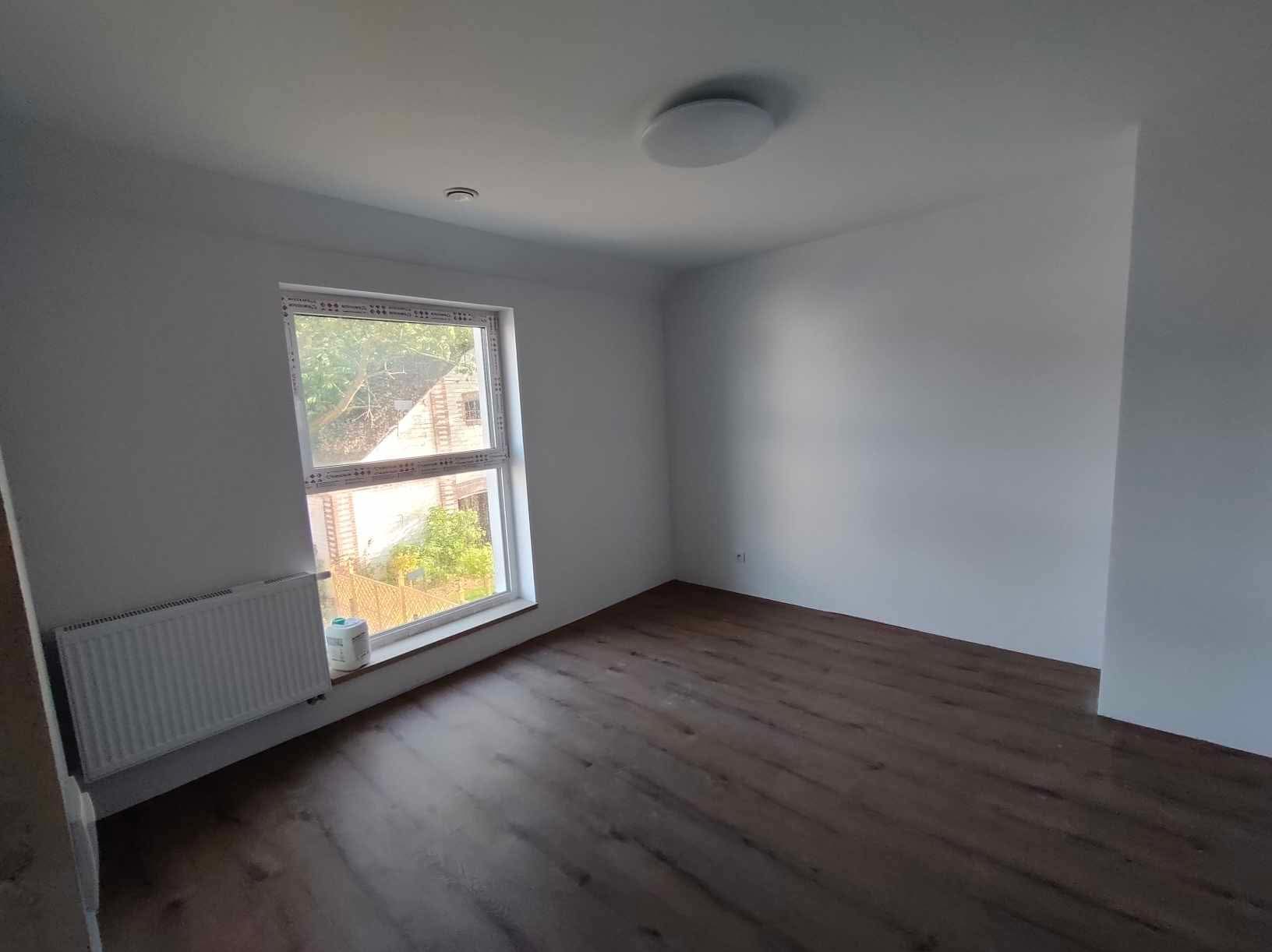 Pusty pokój po remoncie z panelami podłogowymi w odcieniach brązu, białymi ścianami, grzejnikiem pod oknem i okrągłą lampą sufitową.