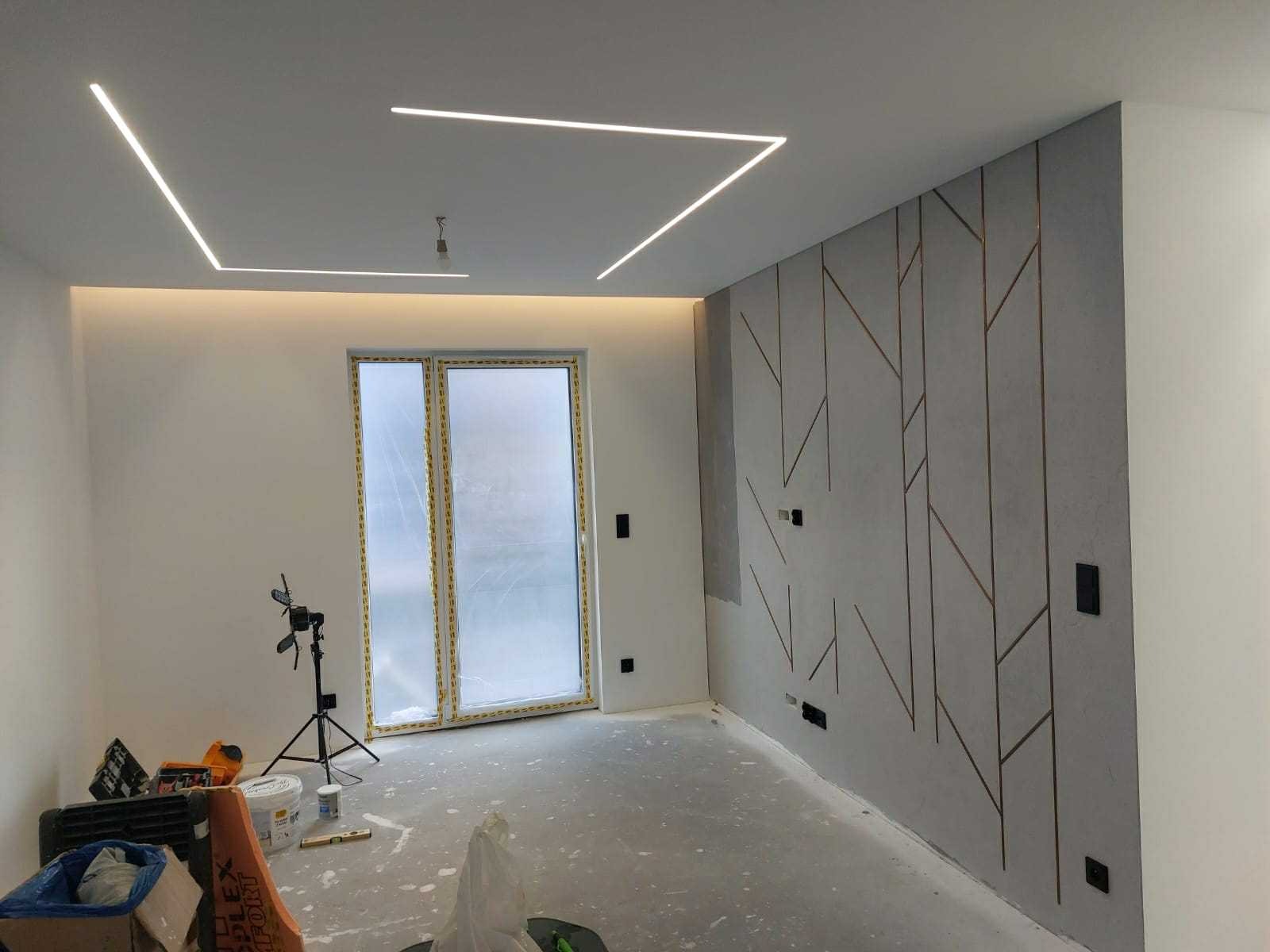 Wnętrze w trakcie wykańczania: szara ściana z geometrycznym wzorem, ukryte oświetlenie sufitowe, okno zabezpieczone folią i narzędzia budowlane na podłodze.