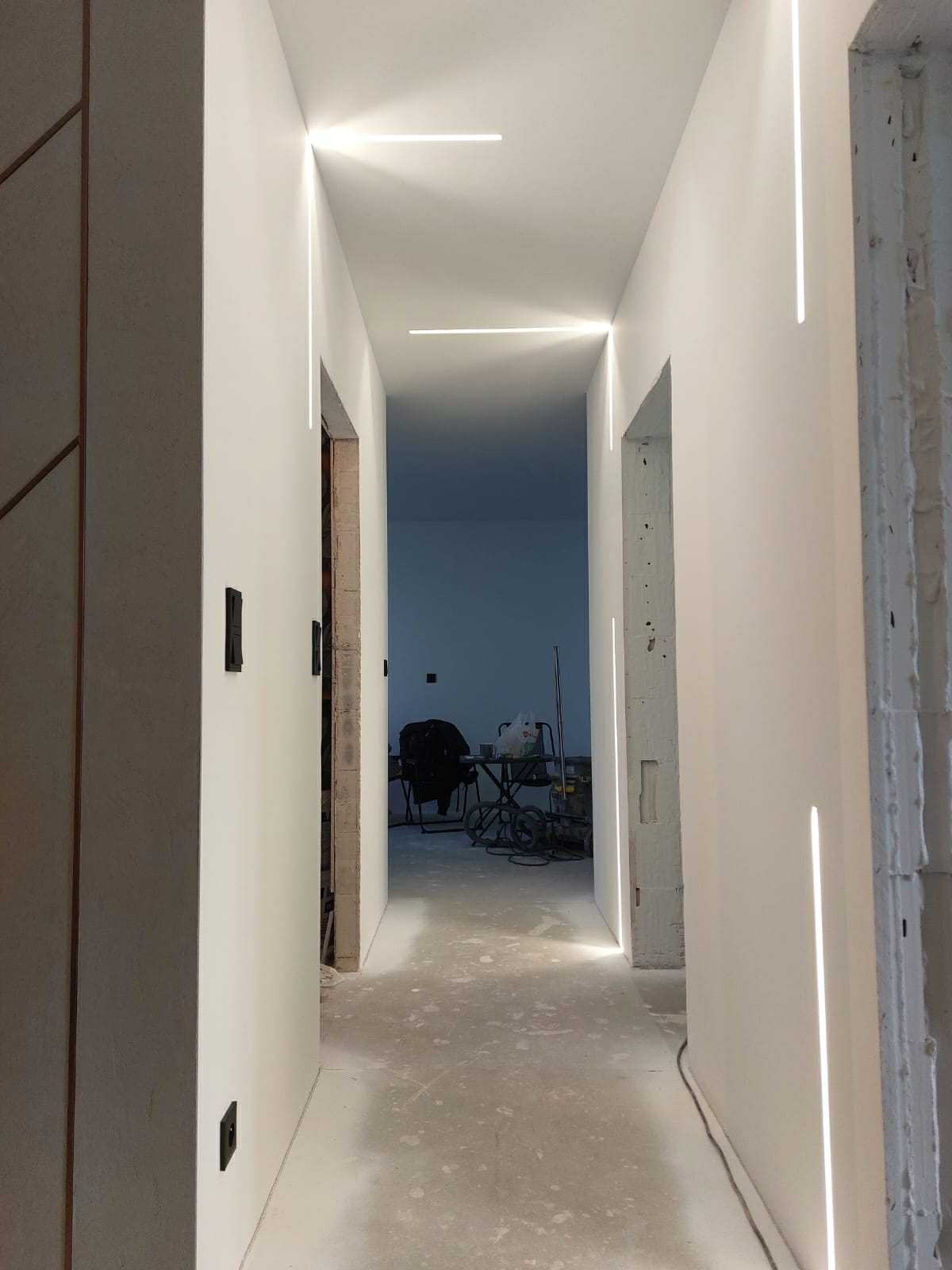 Hallway with modern, linear lighting integrated into the walls and ceiling, freshly painted white, doors without frames, visible tools in the background.
