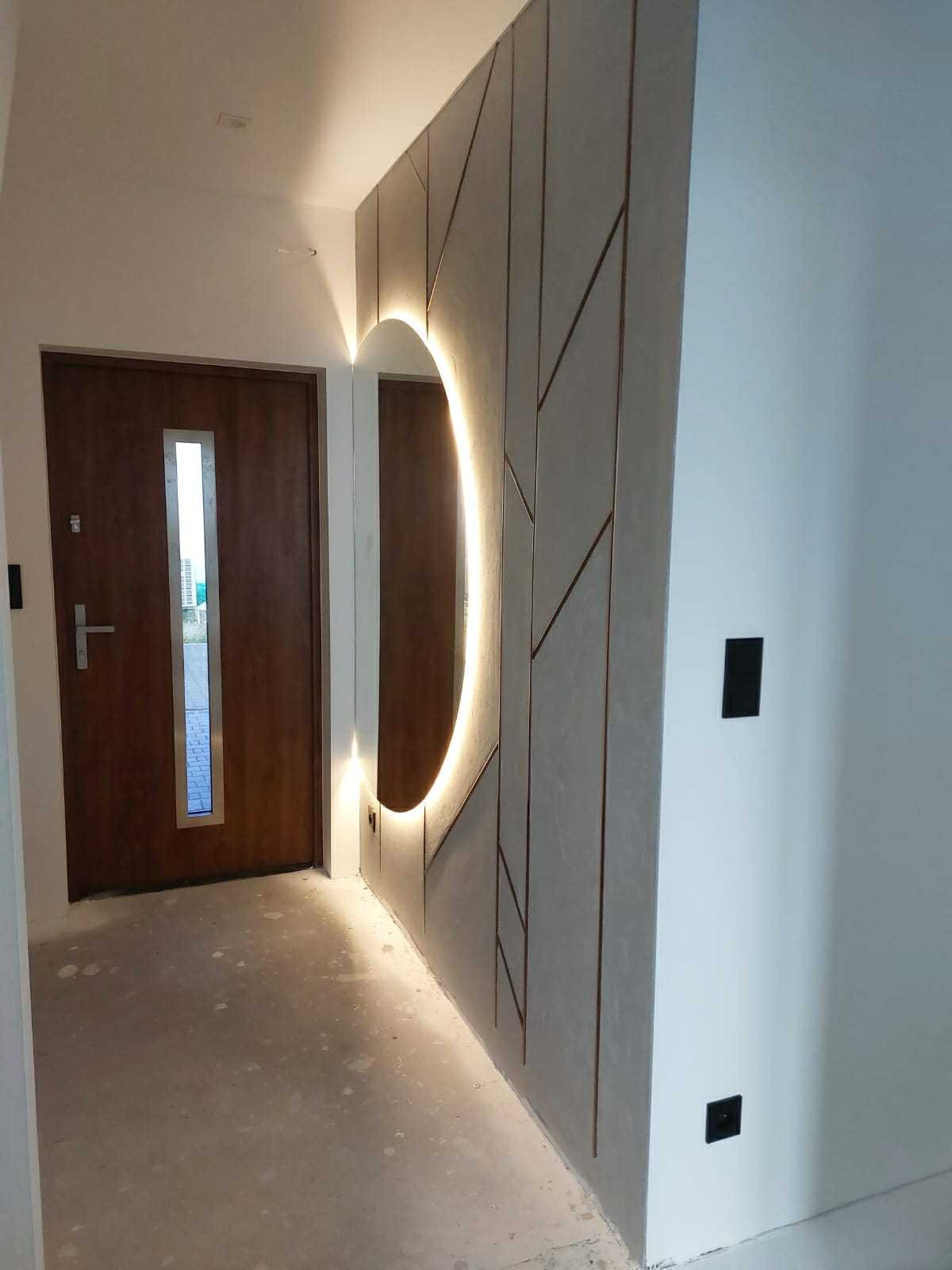 Nowoczesne wykończenie przedpokoju z podświetlanym lustrem i dekoracyjną ścianą. Brązowe drzwi wejściowe z przeszkleniem. Szary surowy beton na podłodze.