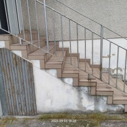J-B.R.W Jakub Wasila - Zewnętrzne schody wyłożone brązowymi płytkami, z metalową balustradą i fragmentem drewnianej osłony z łuszczącą się szarą farbą, przylegające do ściany z widocznymi śladami wilgoci.