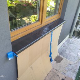 J-B.R.W Jakub Wasila - Montaż czarnych płytek klinkierowych na parapecie zewnętrznym okna z drewnianą ramą, zabezpieczony kartonem i niebieską taśmą malarską, widok z balkonu.