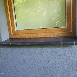 J-B.R.W Jakub Wasila - Okno z drewnianą ramą, osadzone w szarej, teksturowanej elewacji, z ciemnymi, błyszczącymi płytkami parapetu zewnętrznego.