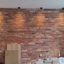 J-B.R.W Jakub Wasila - Ściana z czerwonej cegły z czarnymi reflektorami sufitowymi, widoczne płyty kartonowo-gipsowe i materiały budowlane na podłodze, częściowo wykończone wnętrze.