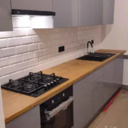 Szare, lakierowane szafki kuchenne wiszące i stojące, drewniany blat z czarnym zlewem i płytą gazową, białe płytki na ścianie, piekarnik w folii ochronnej, podłoga w trakcie wykończenia.