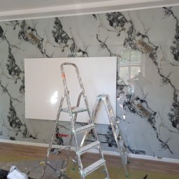 J-B.R.W Jakub Wasila - Ściana wykończona płytami imitującymi marmur z białym panelem, dwie drabiny malarskie, widoczne gniazdka elektryczne. Prace wykończeniowe w trakcie realizacji.