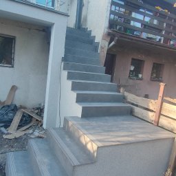 J-B.R.W Jakub Wasila - Zewnętrzne schody z szarych płytek prowadzące do domu, z widocznym wykończeniem elewacji i balustradą balkonu. Ujęcie z dołu, podkreślające solidność konstrukcji.
