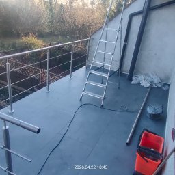 J-B.R.W Jakub Wasila - Balkon z nową, metalową balustradą i szarą podłogą z płytek. Widoczna drabina, narzędzia i otoczenie budynku z zielenią. Ujęcie z góry, w dziennym świetle.