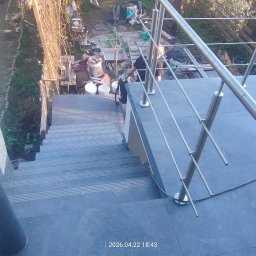 J-B.R.W Jakub Wasila - Zewnętrzne schody z szarych płytek z metalową balustradą. Widok z góry na dół, w tle widoczny ogród z elementami budowlanymi.