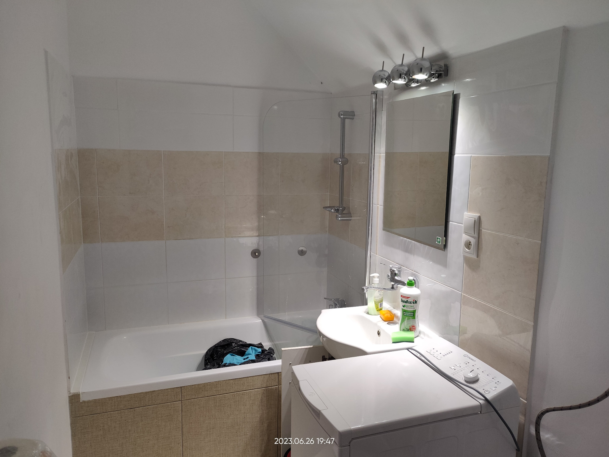 Umywalka z baterią i płynem do naczyń obok pralki w łazience z wanną i ścianami wyłożonymi płytkami w odcieniach bieli i beżu.