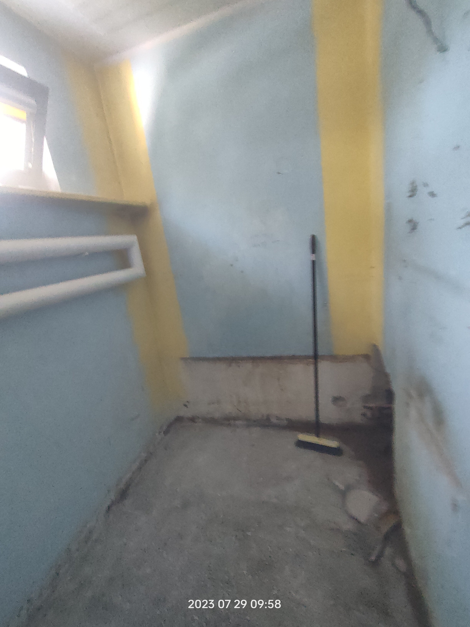 Wnętrze łazienki w trakcie przygotowania do remontu, widoczne stare rury, ściany pomalowane na niebiesko z żółtymi pasami, na podłodze leżą resztki gruzu, w rogu stoi miotła