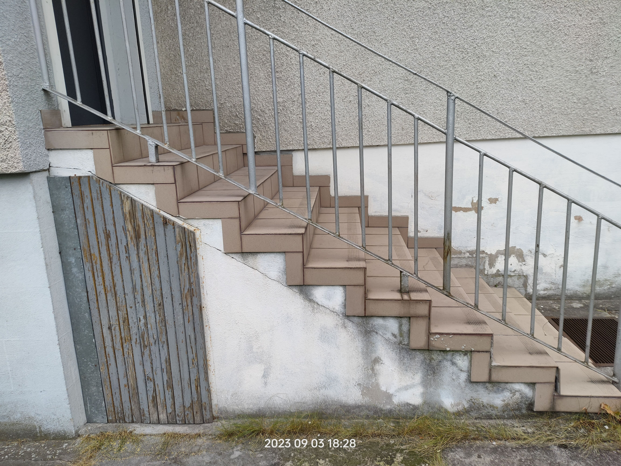 Zewnętrzne schody wyłożone brązowymi płytkami, z metalową balustradą i fragmentem drewnianej osłony z łuszczącą się szarą farbą, przylegające do ściany z widocznymi śladami wilgoci.