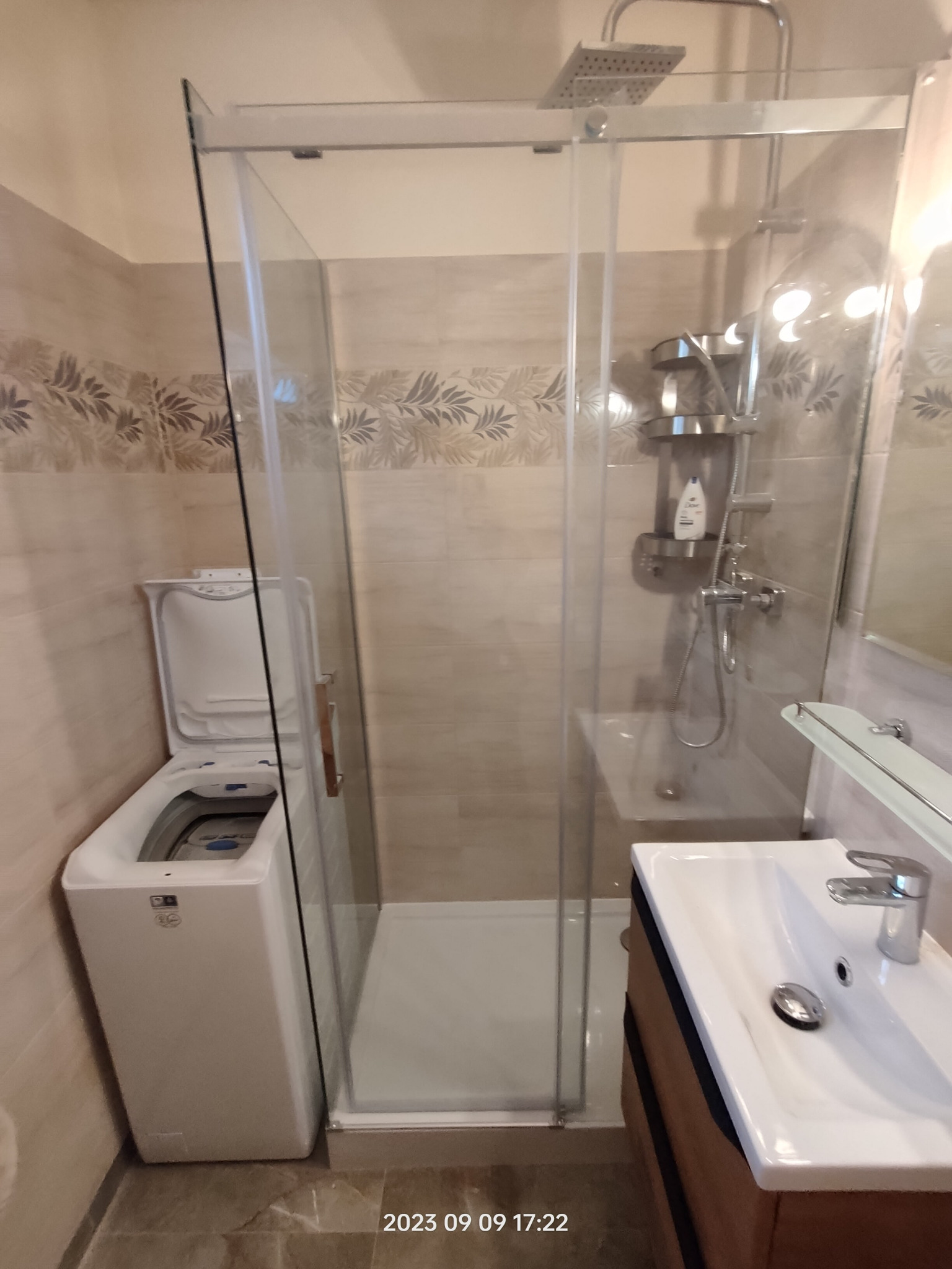 Nowoczesna łazienka z kabiną prysznicową typu walk-in, umywalką z szafką w kolorze drewna i pralką z otwartą klapą, wykończona płytkami z motywem liści.