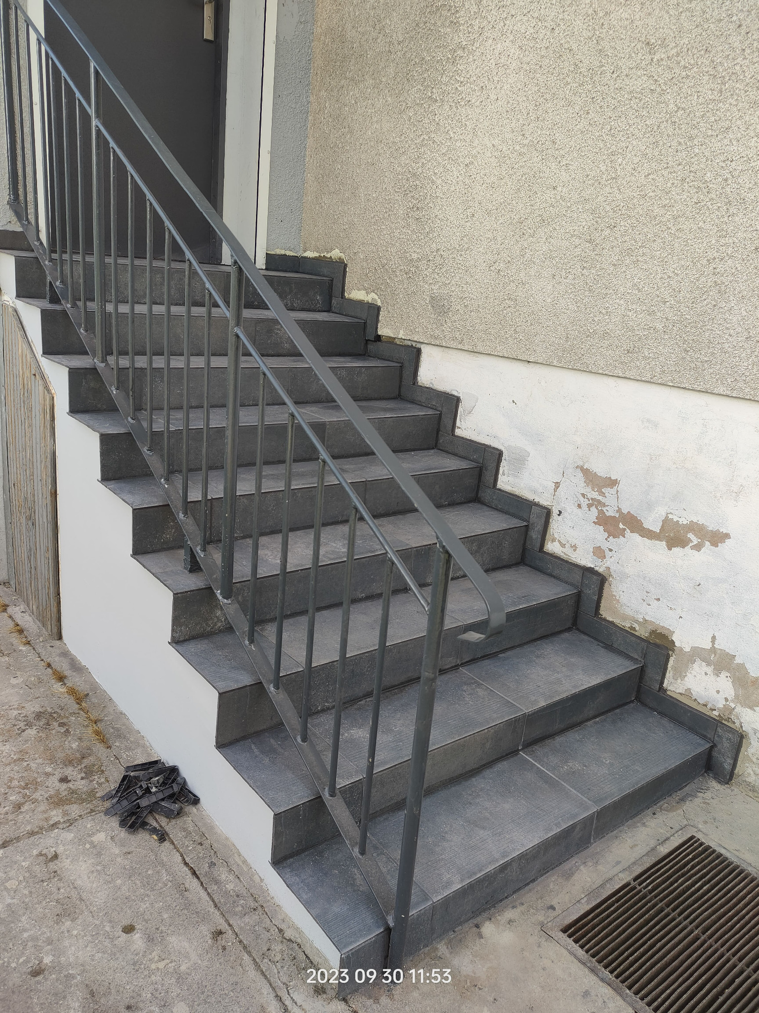 Zewnętrzne schody wyłożone szarymi płytkami z metalową barierką, ściana z widocznymi śladami napraw i odpadającym tynkiem, na dole leżą czarne elementy montażowe.