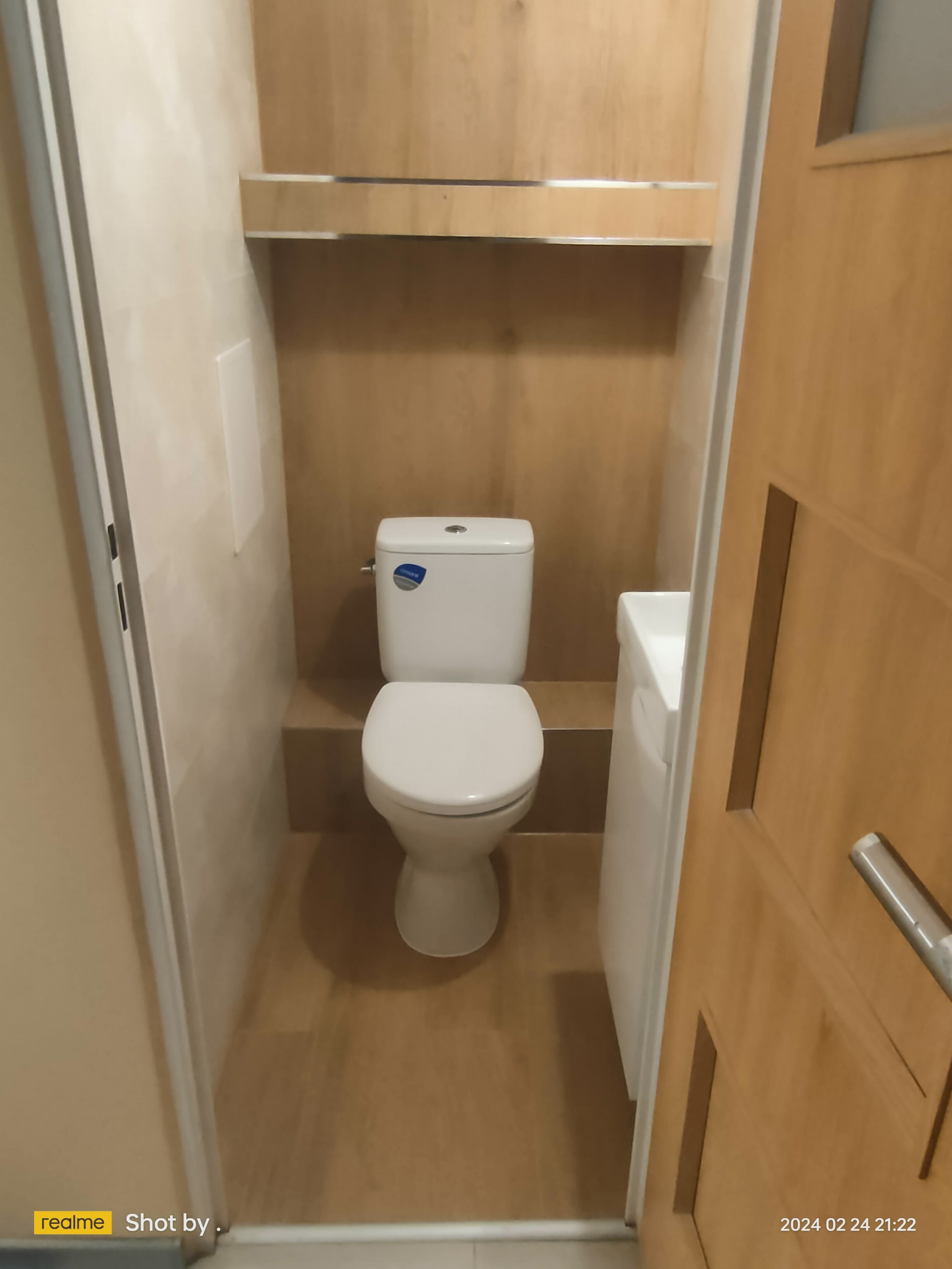 Wnętrze małej toalety z drewnianymi panelami ściennymi i podłogą, widok na muszlę klozetową, umywalkę i częściowo otwarte drewniane drzwi.