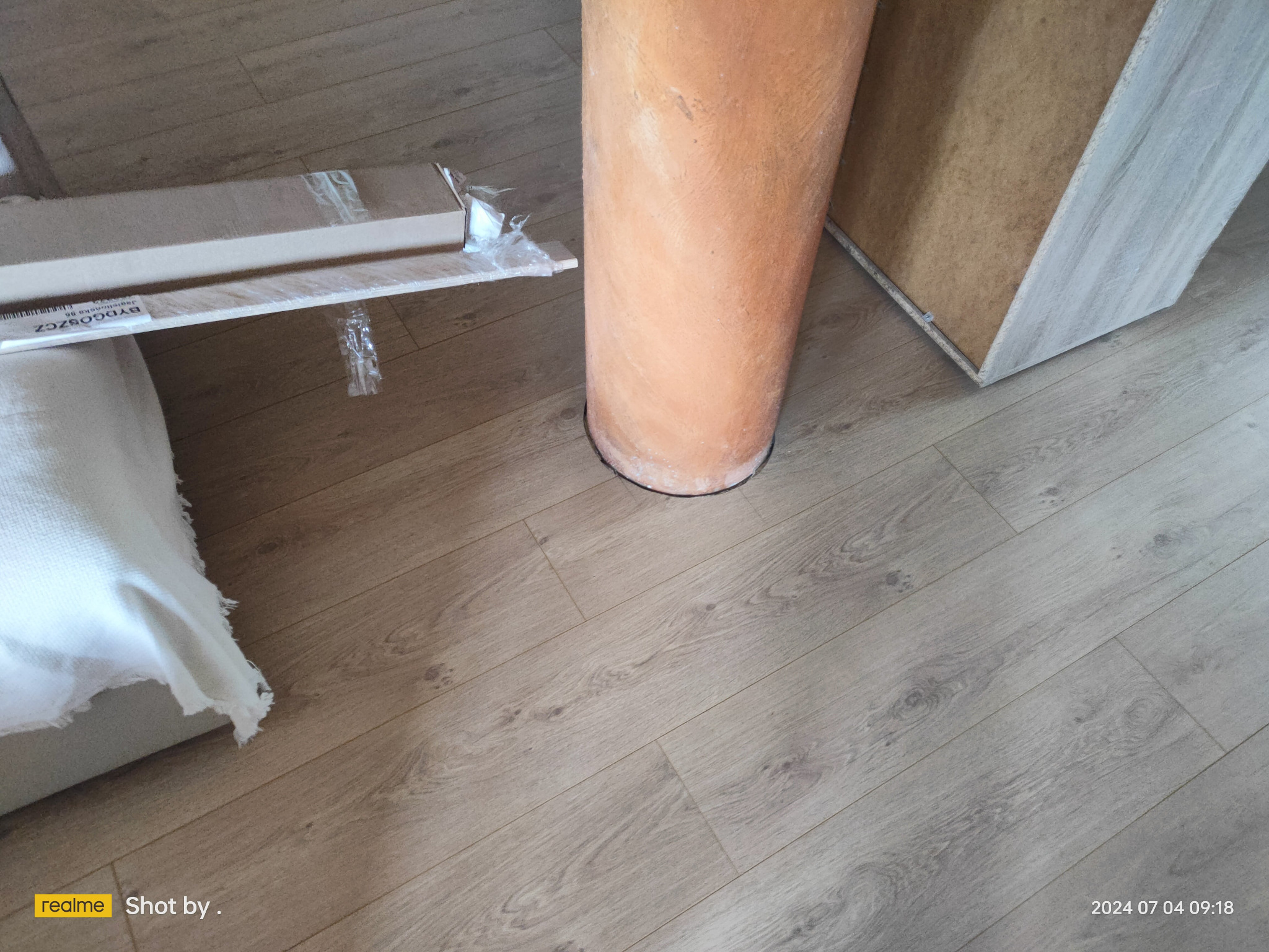 Nowe panele podłogowe w opakowaniu, widoczne na podłodze z jasnego laminatu, obok pionowa rura i fragment mebla, przygotowane do montażu.