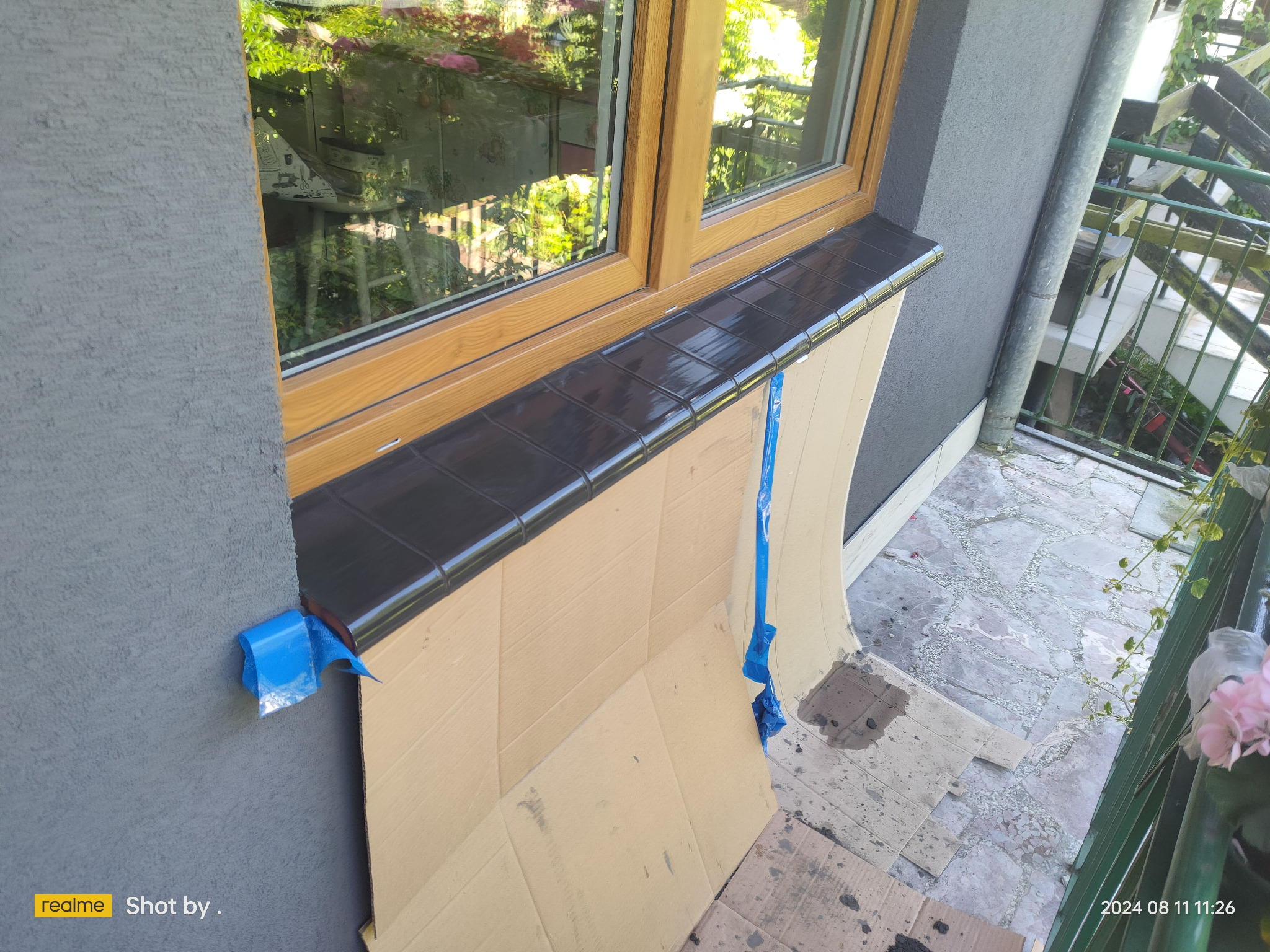 Montaż czarnych płytek klinkierowych na parapecie zewnętrznym okna z drewnianą ramą, zabezpieczony kartonem i niebieską taśmą malarską, widok z balkonu.