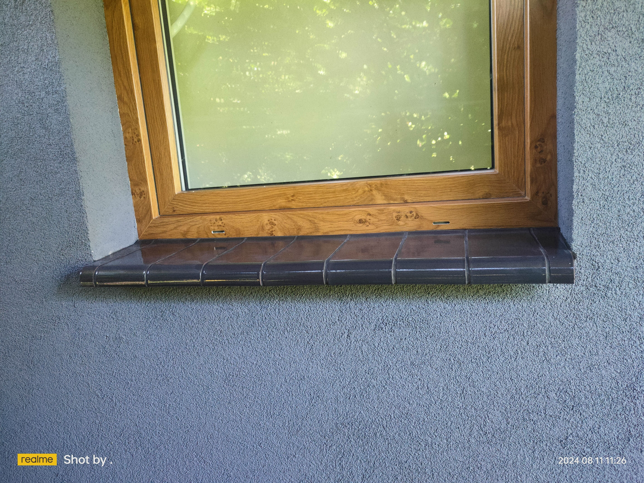 Okno z drewnianą ramą, osadzone w szarej, teksturowanej elewacji, z ciemnymi, błyszczącymi płytkami parapetu zewnętrznego.