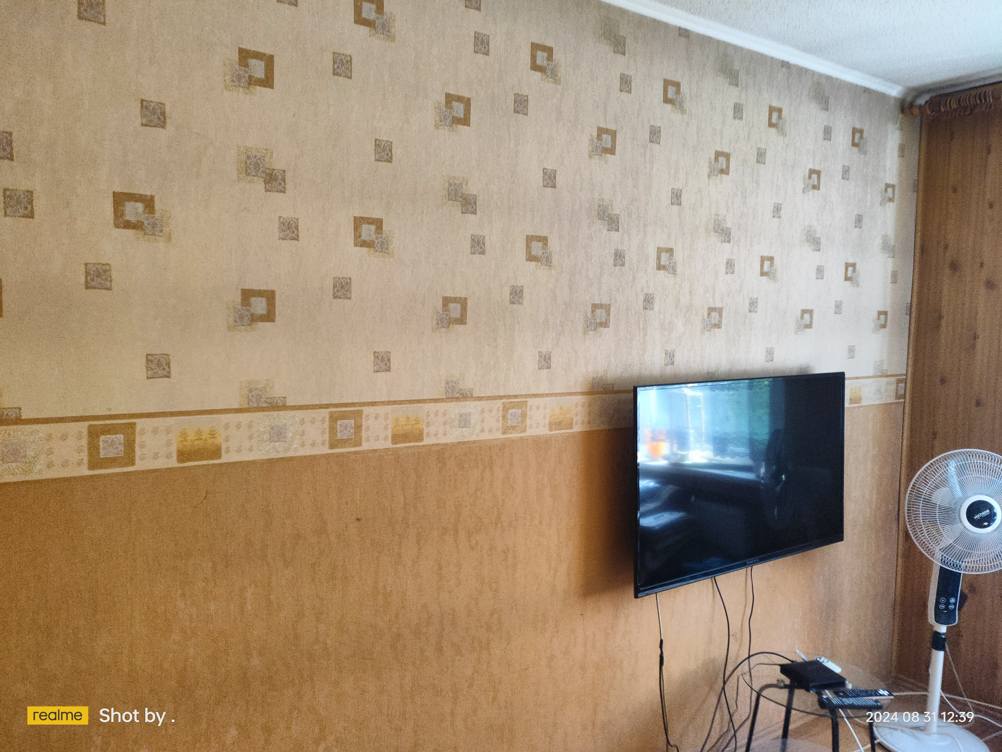 Pokój z dwukolorową ścianą, tapetą w geometryczne wzory w górnej części i telewizorem LCD zawieszonym na ścianie. Widoczny również wentylator stojący.