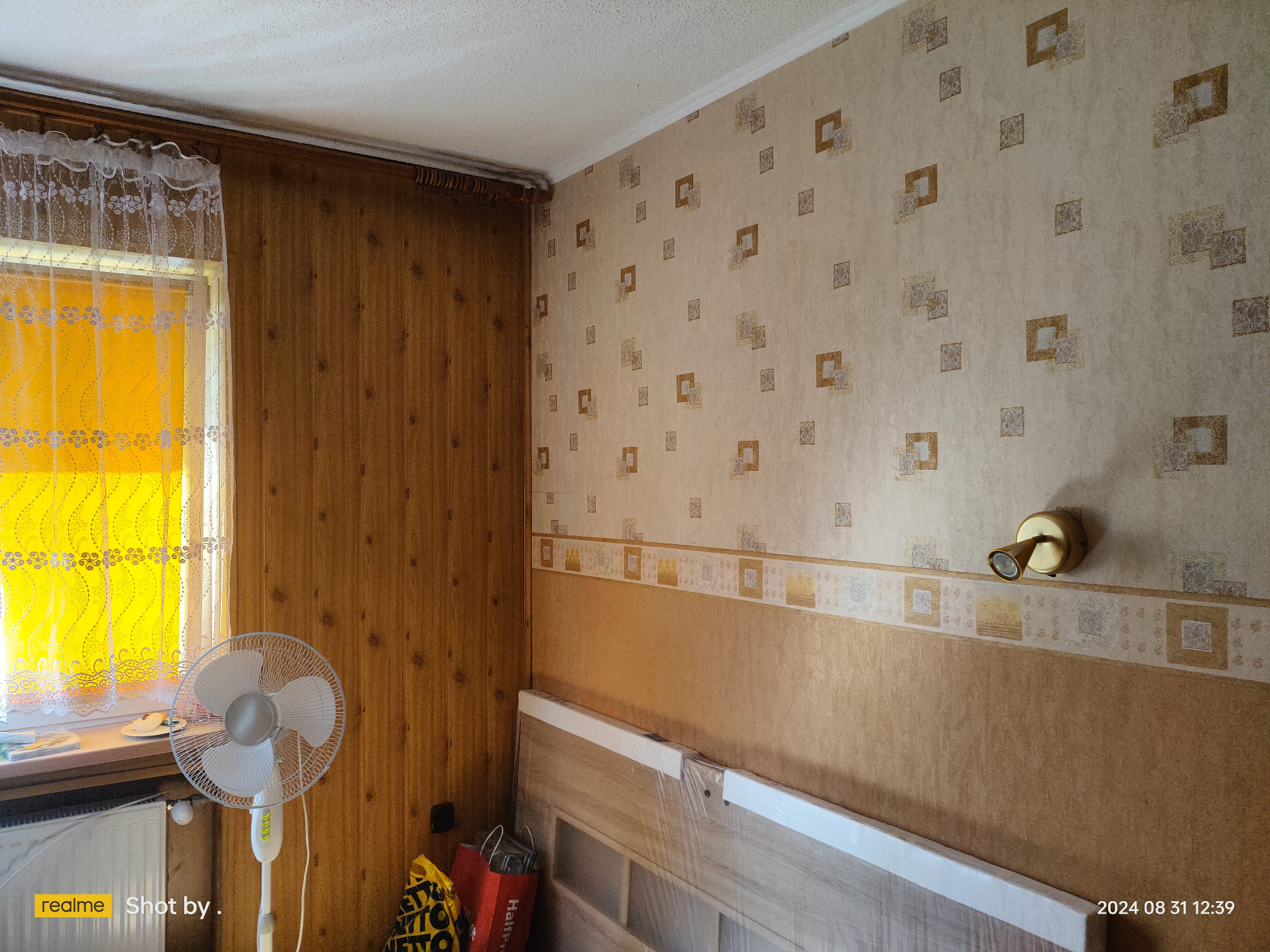 Pokój przed remontem: ściany wyłożone boazerią i tapetą w kwadraty, biały wentylator stojący, okno z firanką, torby na podłodze.