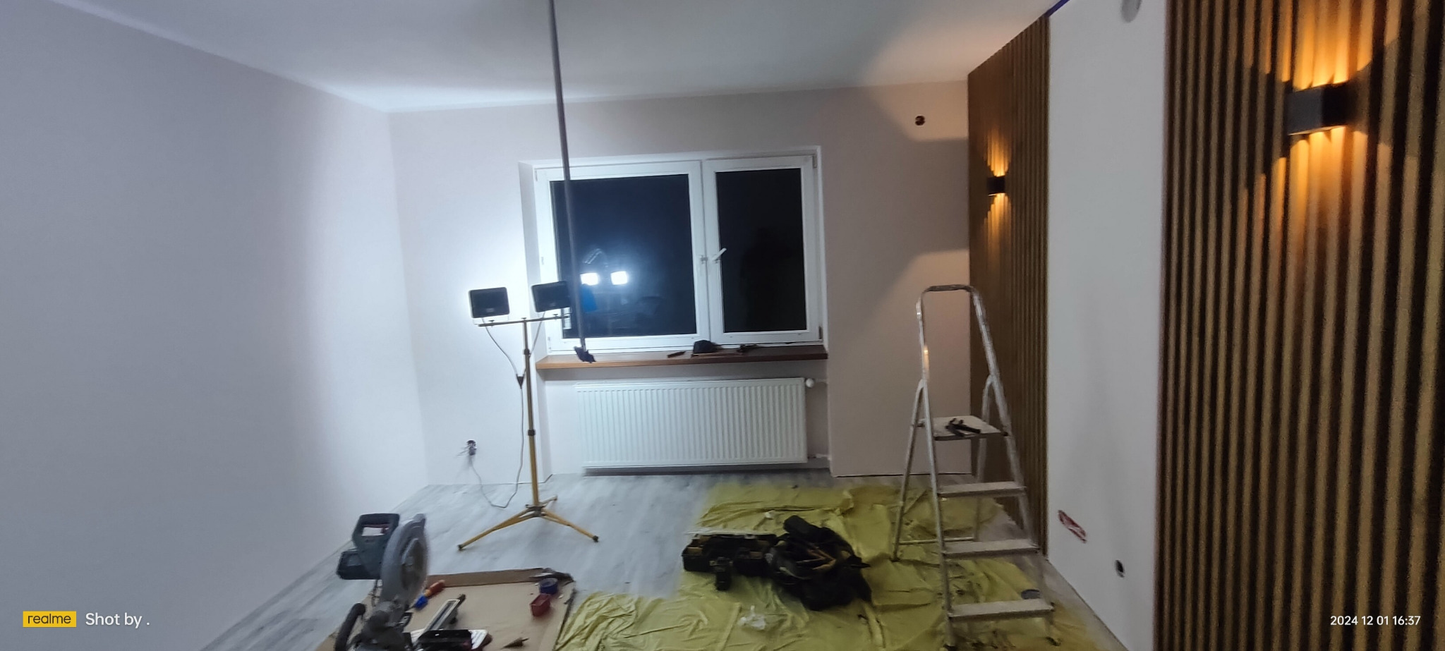 Wnętrze w trakcie remontu: ściany pomalowane na jasny kolor, zamontowane drewniane lamele z oświetleniem, drabina, narzędzia i lampa oświetleniowa na statywie.