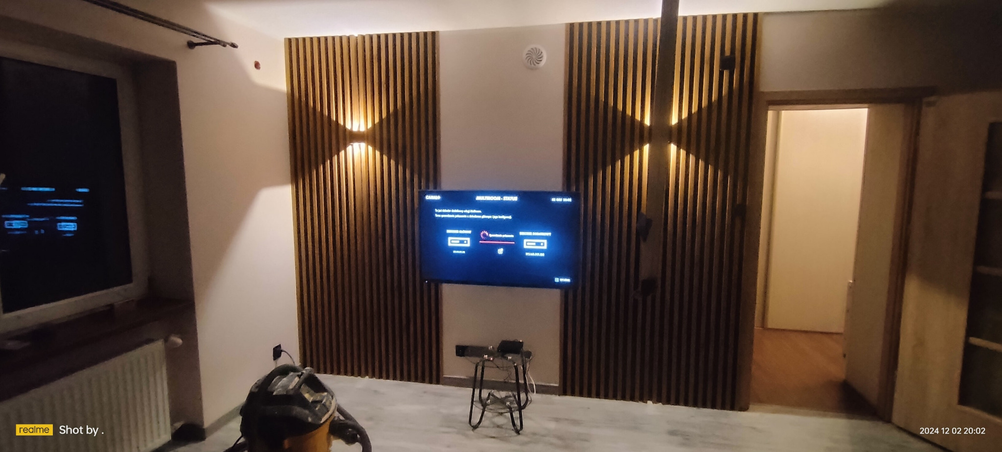 Salon z drewnianymi panelami ściennymi, podświetleniem punktowym i telewizorem na ścianie, widoczny odkurzacz i fragment kaloryfera.