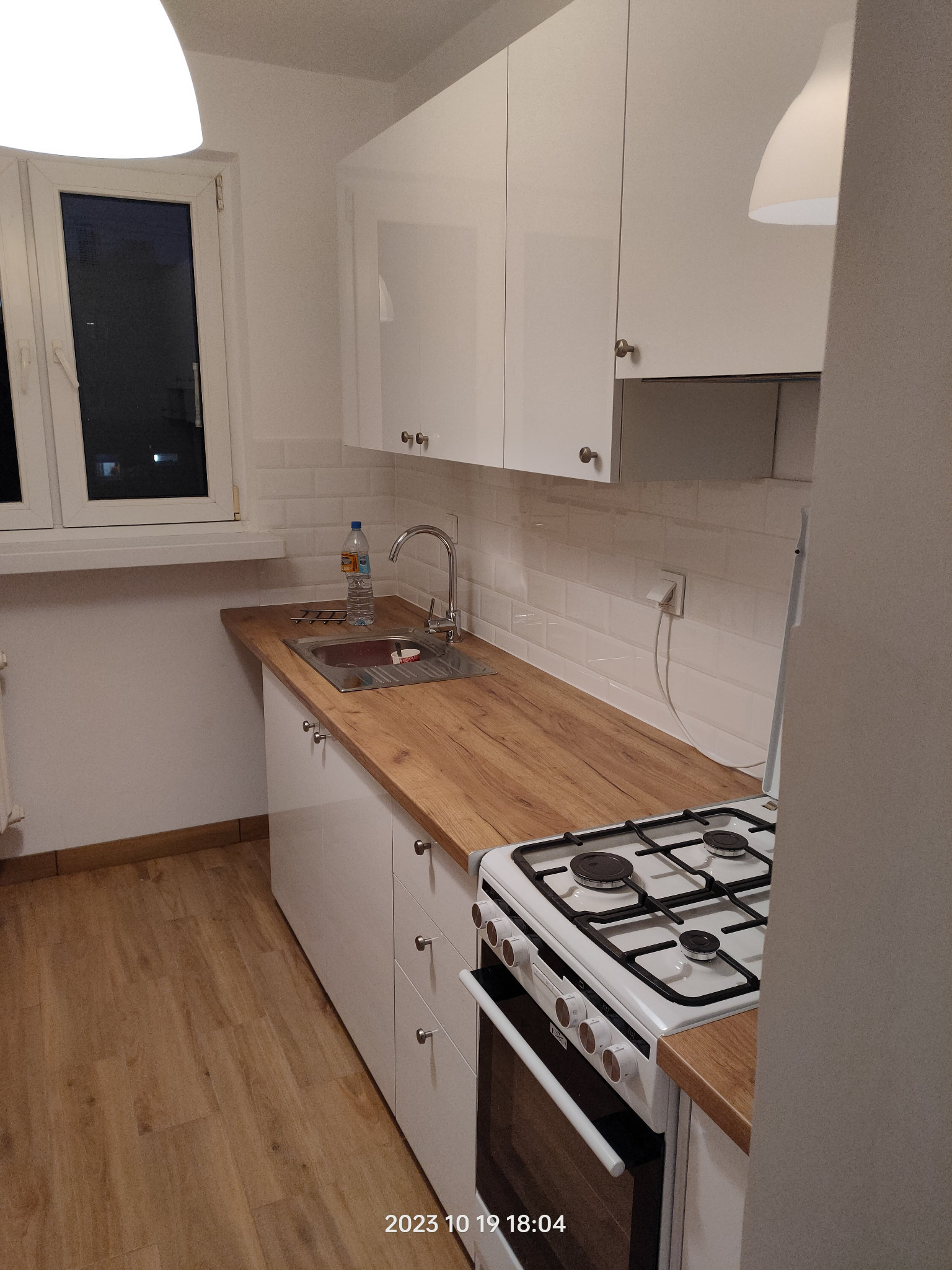 Nowoczesna, jasna kuchnia po renowacji z białymi szafkami, blatem imitującym drewno, białymi płytkami na ścianie i widoczną kuchenką gazową.