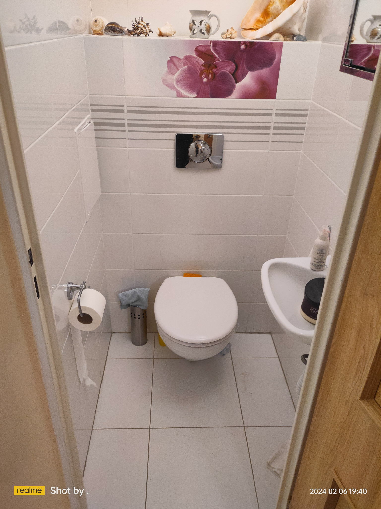 Mała łazienka z białą armaturą, dekoracyjnymi płytkami z motywem orchidei i muszlami na półce, widok z perspektywy drzwi.