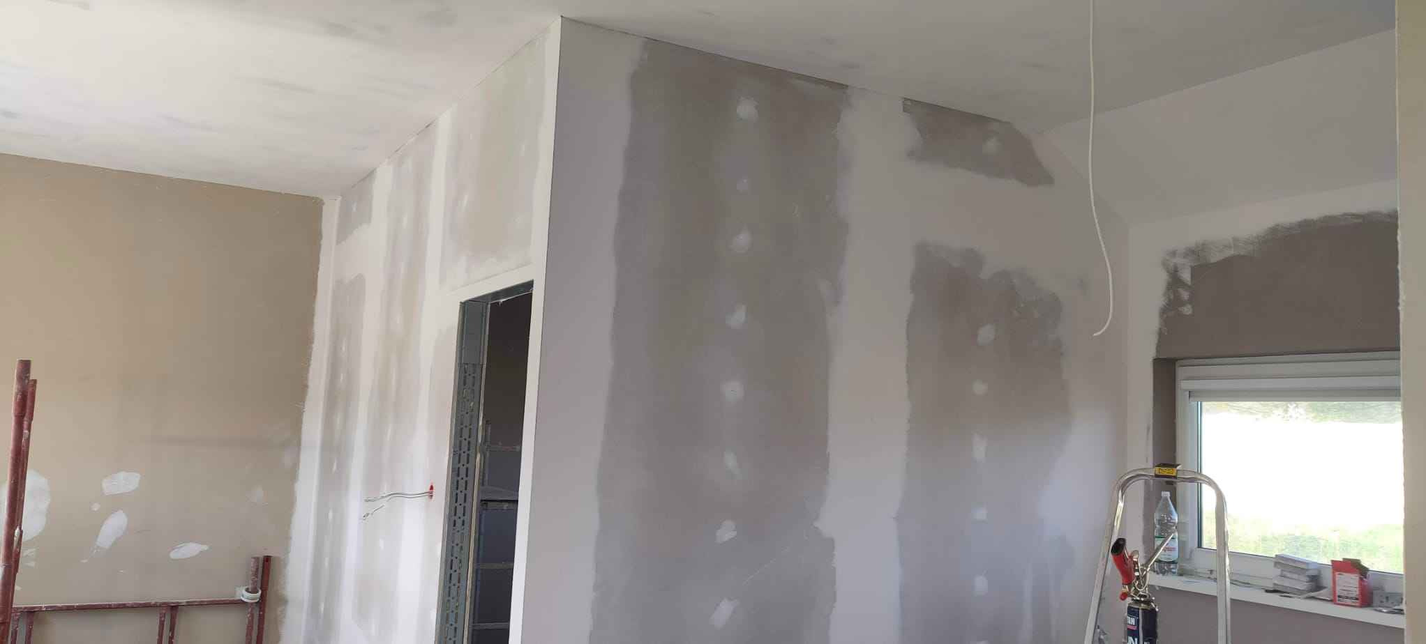 Wnętrze pokoju w trakcie prac wykończeniowych, ściany częściowo pokryte gładzią, widoczne łączenia płyt karton-gips, drabina aluminiowa przy oknie, niepomalowana futryna drzwi.